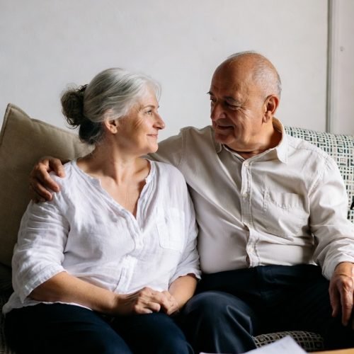 Ontario Wills at home- Elderly couple sitting at home arm in arm with smiles and there are papers on a table infront of them. There is a woman with grey hair in a bun and a bald older man. both are wearing white shirts
